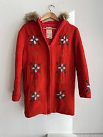 Red wool coat with floral embroidery on front, sleeves, and pockets, hung on a wooden hanger against a white wall. Fur trim on hood visible.