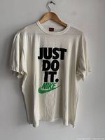 Full front view of white Nike T-shirt with 'JUST DO IT.' text and green swoosh logo on hanger.