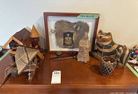 View of entire lot on wooden table showing multiple 3D artwork pieces including the framed shadowbox art, wooden cat sculpture, carved boat, figurines, basket, and stone