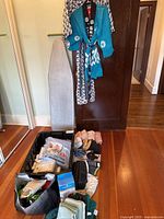 Photo of ironing board, hanging blue and black robe with white symbols, and black bin filled with assorted laundry and bathroom supplies on floor.