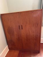 Front view of the teak wardrobe cabinet showing two doors and recessed handles, placed against a corner wall.