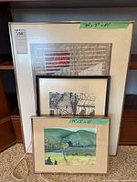 Three framed artworks leaning against a wooden shelf on carpet. The sizes are labeled on the frames as 20 x 27 inches and 14 x 11 inches.
