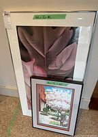 Two framed artworks standing on the floor; taller Georgia O'Keeffe poster behind smaller Robert Amos print, visible green tape size labels