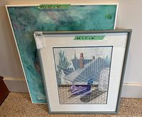 Photo showing both framed artworks leaning against a wall, with size labels visible on tape. Larger piece has abstract blue-green composition, smaller piece depicts rooftops and glass objects with shadow play.