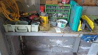 View of the workbench with various plastic organizers containing screws, nails, nails in containers, small hardware parts, a yellow bucket, toolboxes, extension cords coiled on left side.