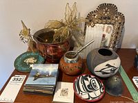 Display of assorted decorative items including pottery, glass candle holders, carved container with metal lid, metallic leaves, postcards and small bell.