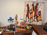 Photo of the international themed artwork collection showing a large fabric painting with stylized figures, wooden sculptures, fabric art, and various decorative items arranged on a wooden table.