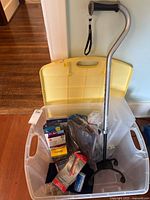 Clear plastic tote with yellow lid containing assorted medical aids. A quad base silver walking cane with black handle is leaning against the tote. Various smaller items are inside the tote in their packaging.