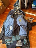 Blue and black vest with Rotary emblem sitting on top of black jackets and other clothing items, shoes to the right side on wooden floor.