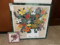 Photo showing two tile mosaic wall hangings, one large square piece with green framing and one smaller square piece with pink framing, both depicting colorful flowers.