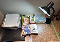 Overview photo showing desk lamp, stack of greeting cards, scrapbook album, loose papers, and plastic organizer box on a desk surface.