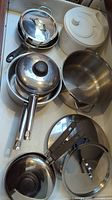 Photo showing several stainless steel saucepans with lids and a white plastic salad spinner in a drawer.