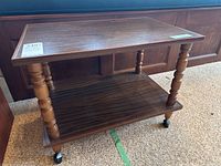 Front and side angled view of the wooden side table showing top and bottom shelves, turned legs, and caster wheels.