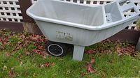 Side and upper view of light gray resin wheelbarrow showing basin and wheels on grassy ground near fence.