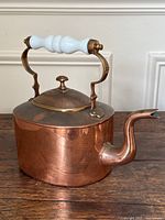Side view of antique copper kettle showing spout, hinged lid, and white milk glass handle with brass fittings.