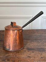 Copper pot displayed on wooden surface at angle showing body, lid, long handle, and pouring spout.