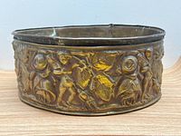 Front view of round antique brass planter showing embossed cherubs and leaves design with some wear