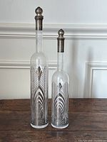 Two tall slender glass bottles with silver plated Art Deco style geometric designs and matching silver plated stoppers, varying heights, placed on wooden surface against white wall.