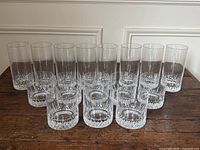 Eight tall highball glasses in back row and eight short whisky tumblers in front row on wooden surface showing matching cut-crystal pattern