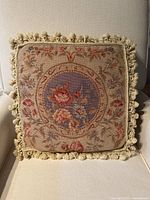 Front view of square needlepoint floral pillow with detailed circular flower design in blue, pink and beige, edged with cotton fringe.