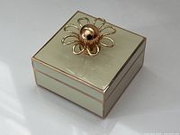 Top and angled side view showing cream enamel box with gold-tone floral handle and metal framing.