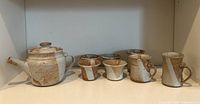 Photo showing full set of teapot, 4 small cups, 2 medium mugs, and 1 taller mug with brown and grey speckled glaze pottery against white background.