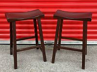 Two Pottery Barn wooden bar stools with dark finish, curved seats, and simple straight legs against a red metal door background.