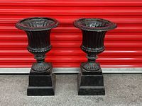 Front view of both black cast iron urn planters on pedestals against red background