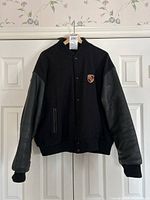 Full front view of black varsity jacket hung on hanger showing wool body, leather sleeves, ribbed hem and snap button closure