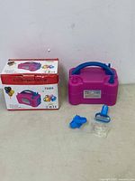 Purple electric balloon pump with blue handle and attached label, shown with three blue/clear nozzles and original product box, front angle.