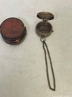 Open brass pocket compass with chain shown next to the round leather case on a white surface.