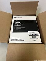 Open cardboard box showing a pack of Grand & Toy white labels with details: permanent adhesive, for mono lasers, 4 x 1 inch size, 2000 labels, compatible with laser, inkjet, copier.