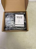 Box opened showing 12 black heavy duty steel hooks, plastic wall anchors and metal screws in small bags, with an installation instruction manual on top.