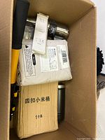 Top view inside a cardboard box containing mixed household and kitchen items including a stainless steel vacuum flask, a boxed item with foreign text, wrapped items, and a yellow handled tool.
