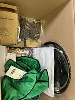 Box contents showing green fabric item with white ribbon, white and green fabric pieces, black belts or straps, brown items in plastic, and small cardboard box visible
