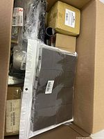 Top view inside the box showing multiple sealed and unsealed smaller boxes, plastic wrapped items and a pack of black material parts labeled with measurements.