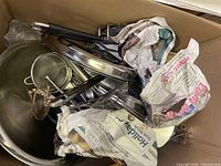 Box with assorted kitchen tools including metal bowls, strainers, spatula, tongs, and items wrapped in newspaper