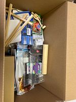 Box containing various craft items such as wooden frames, pens, beading supplies, and paper