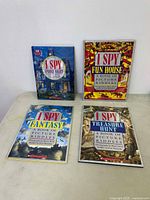 Four I Spy books displayed against a white wall on a table. Titles visible are I Spy Picture Riddles, I Spy Fun House, I Spy Fantasy, and I Spy Treasure Hunt.