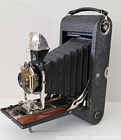 Side angle of antique Kodak bellows camera showing folded bellows, lens, and viewfinder with worn black leather case.