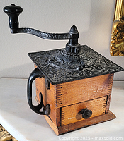 Front angle view of the antique coffee grinder box, showing wooden body and ornate cast iron handle and top.