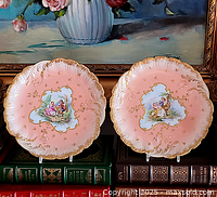 Two pink hand painted Limoges plates with gold detailing and romantic scenes on display standing against a painting.