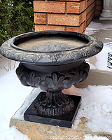 Front side view of large black decorative garden urn showing ornate leaf and floral detailing on rim and body with a square pedestal base; placed against brick wall with snow on ground.
