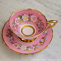 Angled side view of teacup seated on saucer showing pink background, floral design and gold trim