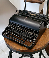 Full side and front view of black Olympia typewriter on small round wooden table showing keyboard and brand name on paper holder.