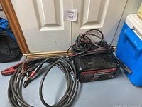 Motomaster battery charger with attached jumper cables on floor in front of white door.