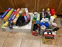 Various cleaning and household supplies including wraps, Ziploc bags, liquid bottles, sprays, and the iRobot Roomba 595 box on floor with tile and wood paneled background.