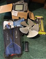 Fish finder sonar depth sounder device taken from box, Rapala fillet knife still in package, life vest in mesh bag, and metal fan shaped object arranged on green carpeted floor.