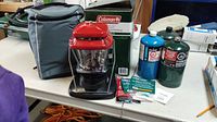 Red and black Coleman propane lantern with clear globe, original green box behind, grey carry case, three propane tanks (blue, green Extra Heavy Duty, black Extra Heavy Duty) on table.