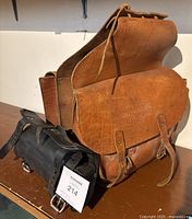 Brown double sided Harley-Davidson leather motorcycle saddlebag and black leather front fork pouch displayed on table, showing embossed logo and buckle straps.